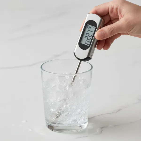 A hand inserting a meat thermometer probe into a glass of ice water for calibration.